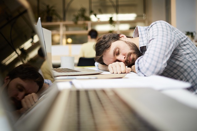 A man with a beard is asleep at his desk, his head resting on his arm, next to a laptop and scattered papers in an office environment—perhaps dreaming of ways to automate a wide array of customer outreach efforts.