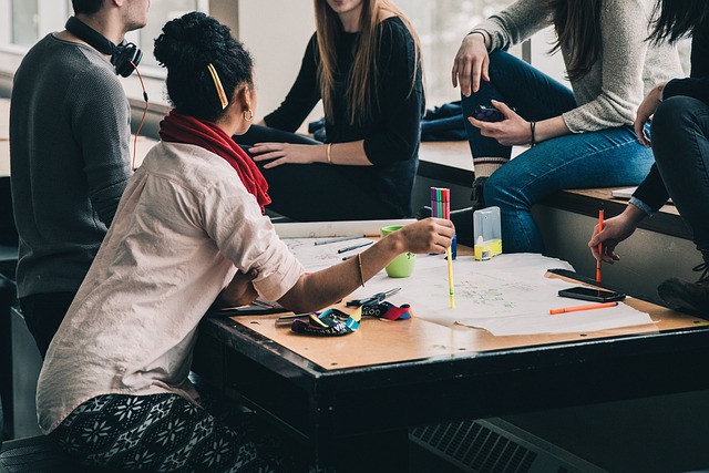 A group of people sit around a wooden table, engaging in conversation and working on various tasks with papers and colored pens spread across the surface.