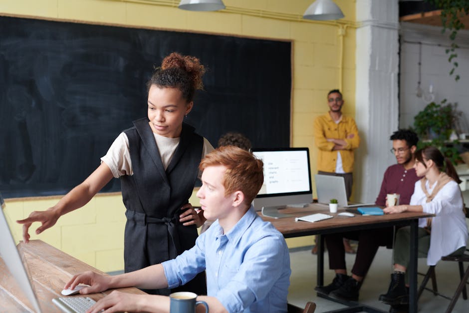 People working in a modern office. A woman points at a computer screen while a man types. Three others are seated in the background, one leaning on a wall.