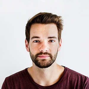 A man with short brown hair and a beard, wearing a maroon shirt, stares directly at the camera against a plain white background.