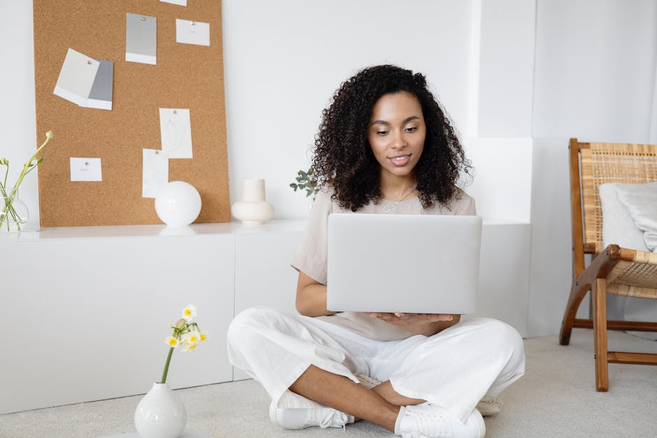 A person with curly hair sits on the floor holding an open laptop, likely exploring affordable web design solutions. In the background, a corkboard with papers, a small vase with flowers, and a wicker chair complete the cozy setting.