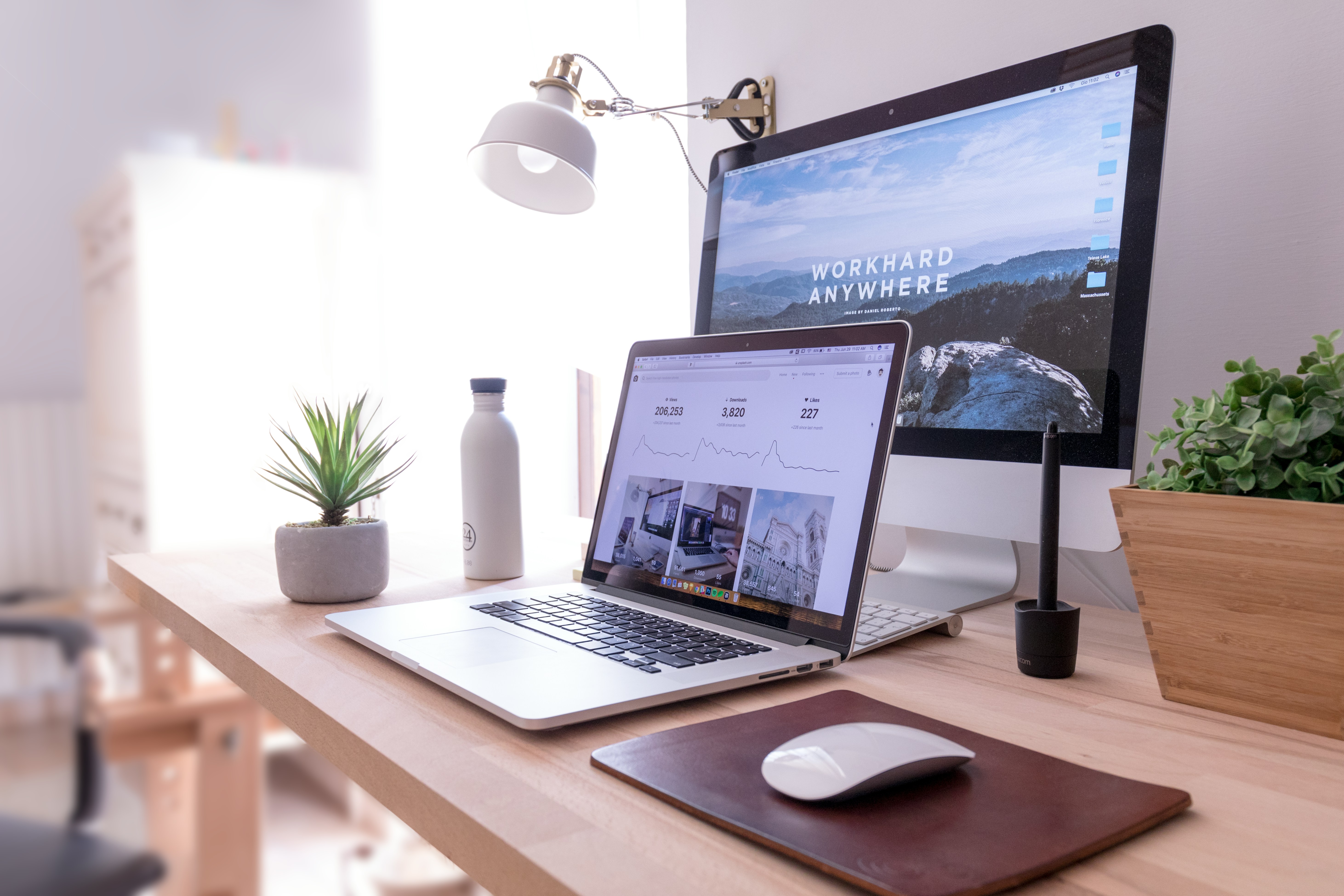 A home office desk setup with a laptop, a desktop monitor displaying Work Hard Anywhere, a potted plant, a water bottle, a graphic tablet, and a mouse on a mouse pad