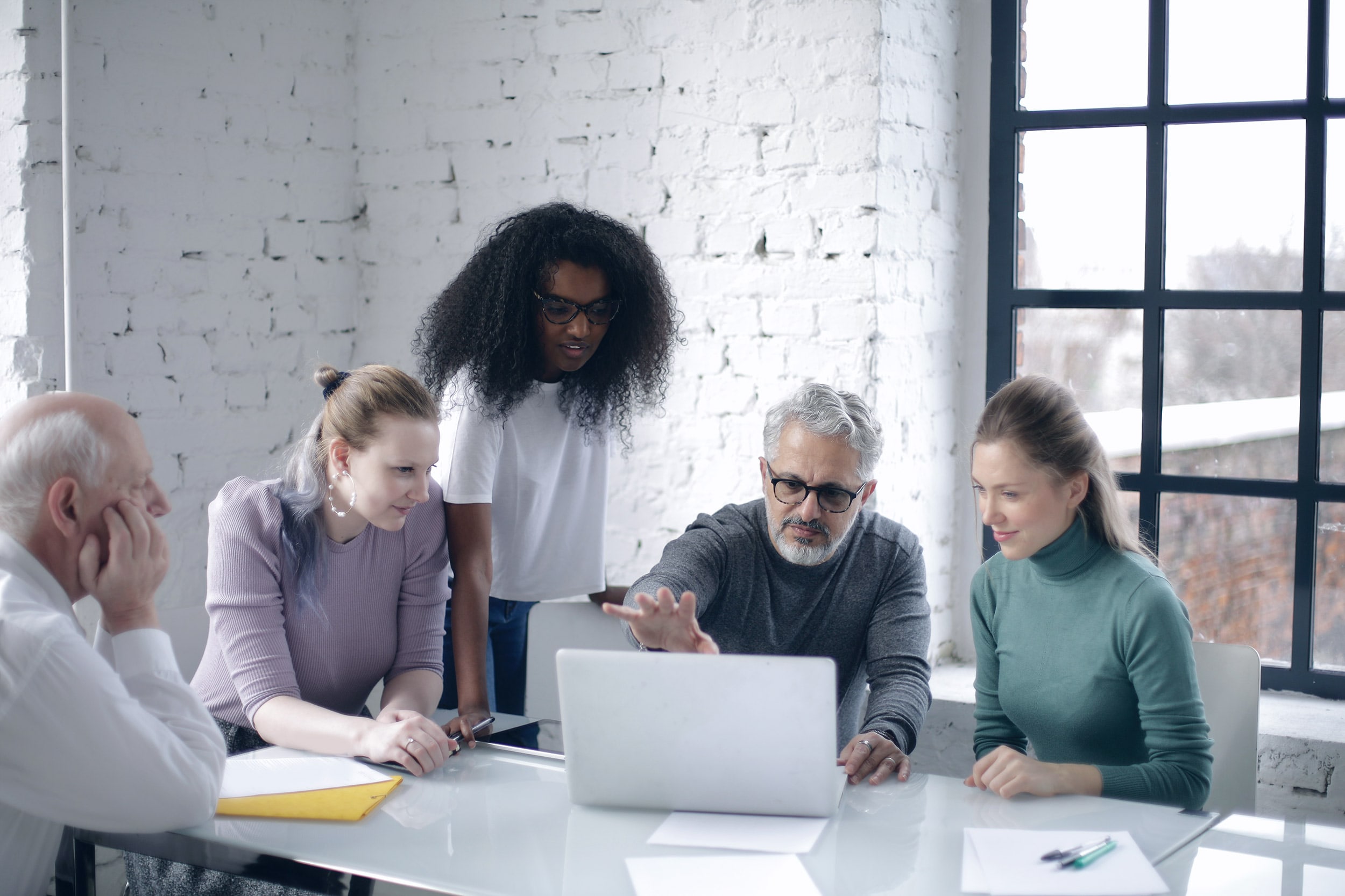 A group of five people gathered around a table, discussing and looking at a laptop screen in a bright room with white brick walls and a large window, brainstorming online marketing strategies for small businesses.