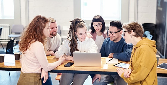 A group of six people gather around a laptop at a wooden table in a bright room, collaborating on social media management and intently looking at the screen.