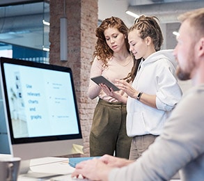 Three individuals are working in an office. Two people are examining a tablet, likely reviewing web design concepts, and another person is sitting at a computer. A monitor displays a presentation.