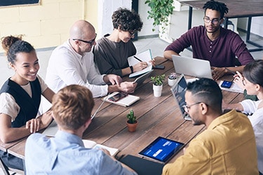 A diverse group of people sit around a wooden table in an office setting, engaged in a meeting. Laptops, tablets, and notebooks are on the table alongside a small plant in the center. They discuss various service requests while ensuring accurate contact information is kept up to date.