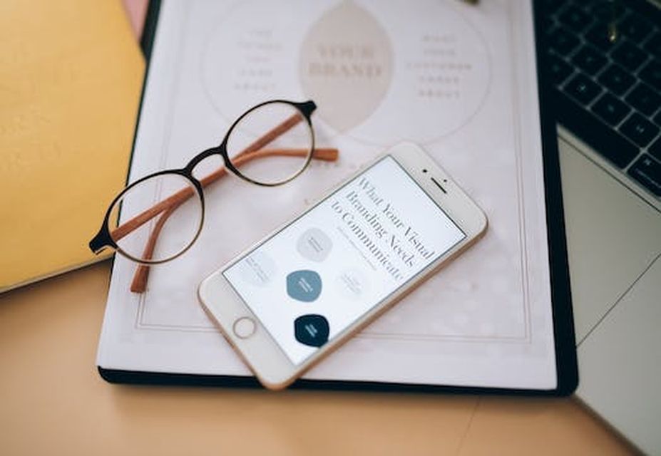 A pair of eyeglasses rests on a folder beside an iPhone displaying a screen with minimalist icons, all placed on a desk near a laptop where affordable web design solutions are being explored.