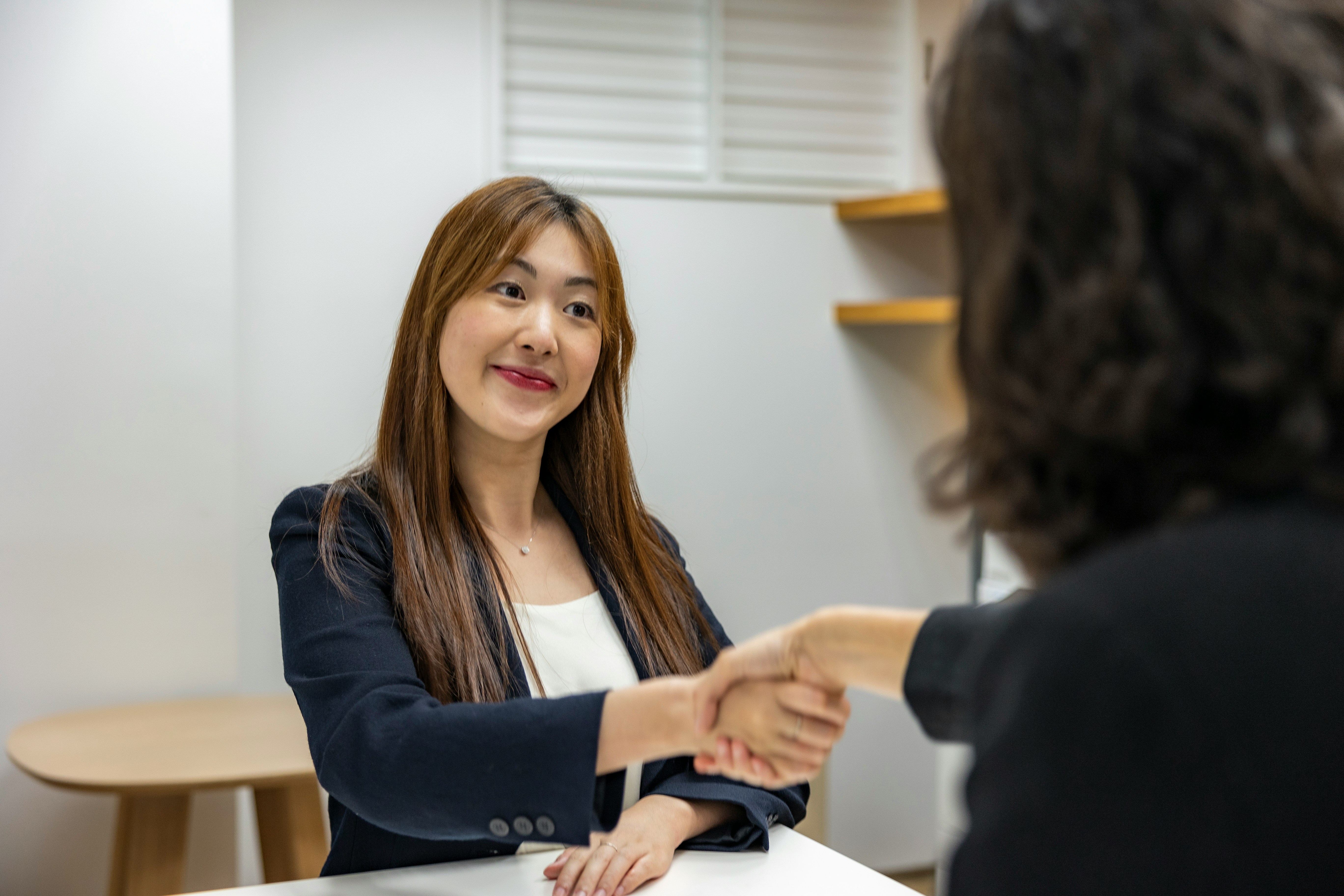 Two women are seated opposite each other at a table, shaking hands. One woman, with long, straight hair and wearing a blazer, appears to be discussing talent attraction strategies. The other woman's face is not visible.