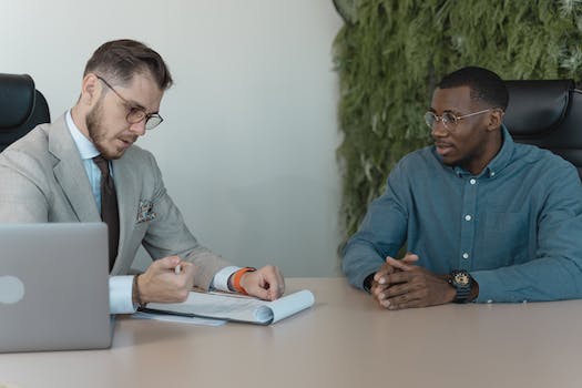 Two men sit at a desk in a meeting room. One man, wearing a suit and glasses, points at a document, while the other, in a casual shirt and glasses, listens attentively. Their discussion covers employer branding strategies to enhance recruitment marketing efforts.