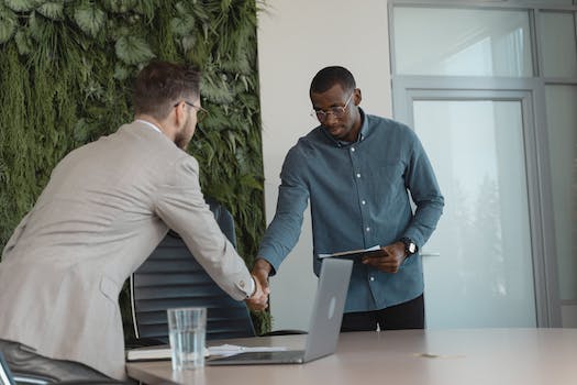 Two men, one in a beige blazer and the other in a blue shirt, are shaking hands in an office setting with a laptop and a glass of water on the table, discussing innovative hiring strategies to strengthen workforce development.