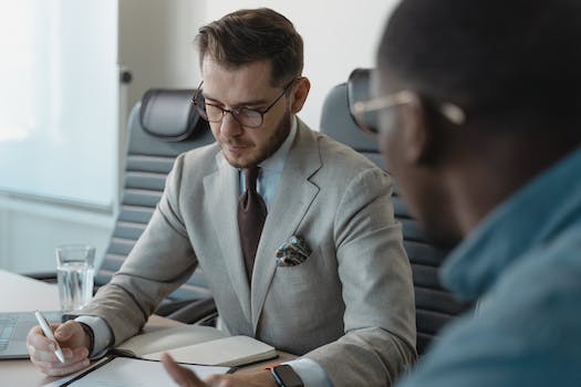 Two men sit at a table in a business setting. The man on the left writes in a notebook, while the man on the right observes him attentively. Both are wearing glasses and formal attire, likely discussing strategies for talent acquisition.