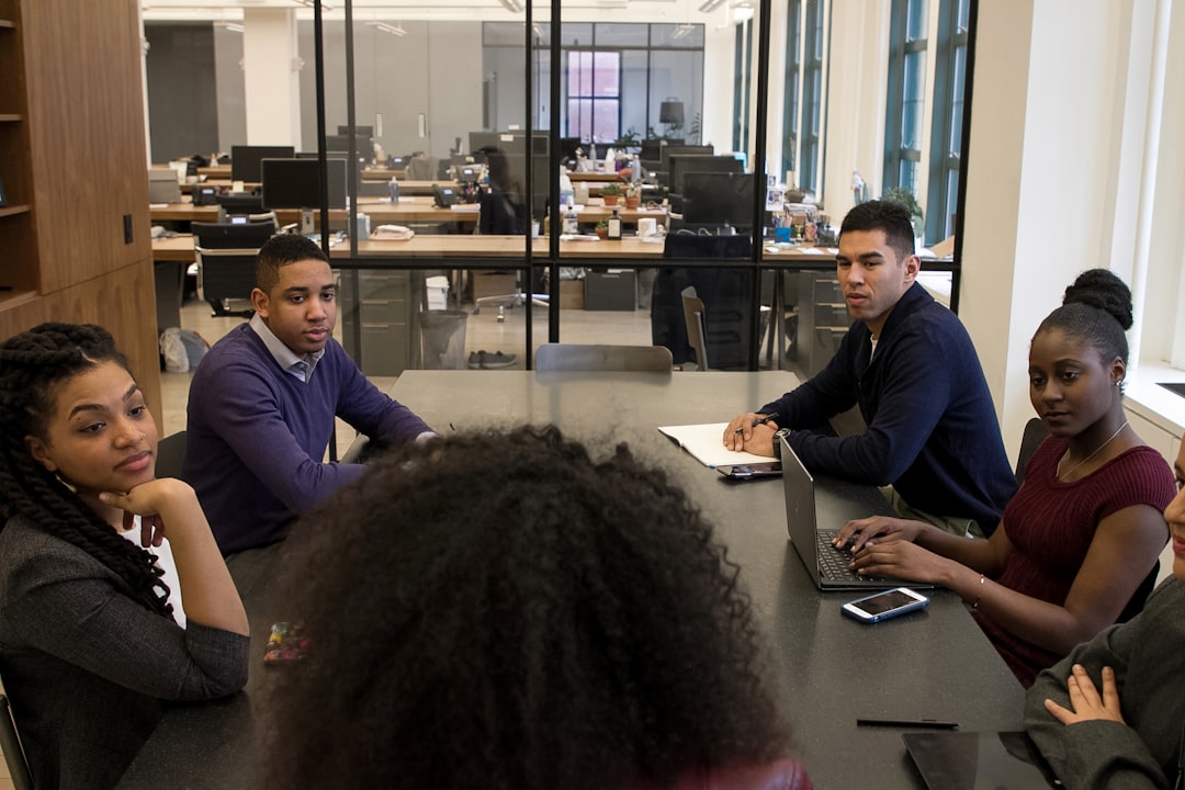 Five people are seated around a table in a conference room, discussing analytics in recruitment. Laptops and other office materials are on the table, underscoring the importance of employer branding. An open office space is visible in the background, emphasizing the collaborative environment.