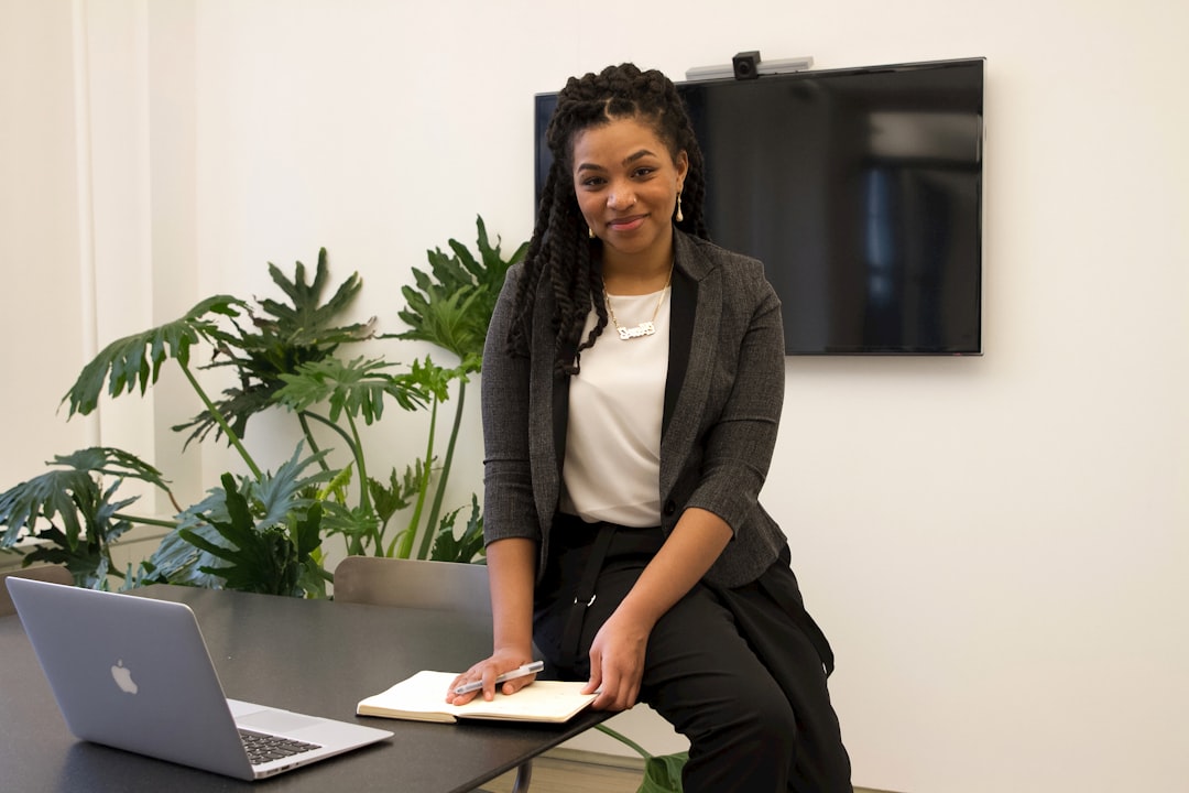 A person with braided hair sits on the edge of a table holding a notebook and pen, with a laptop and large screen in the background, possibly exploring analytics in recruitment.