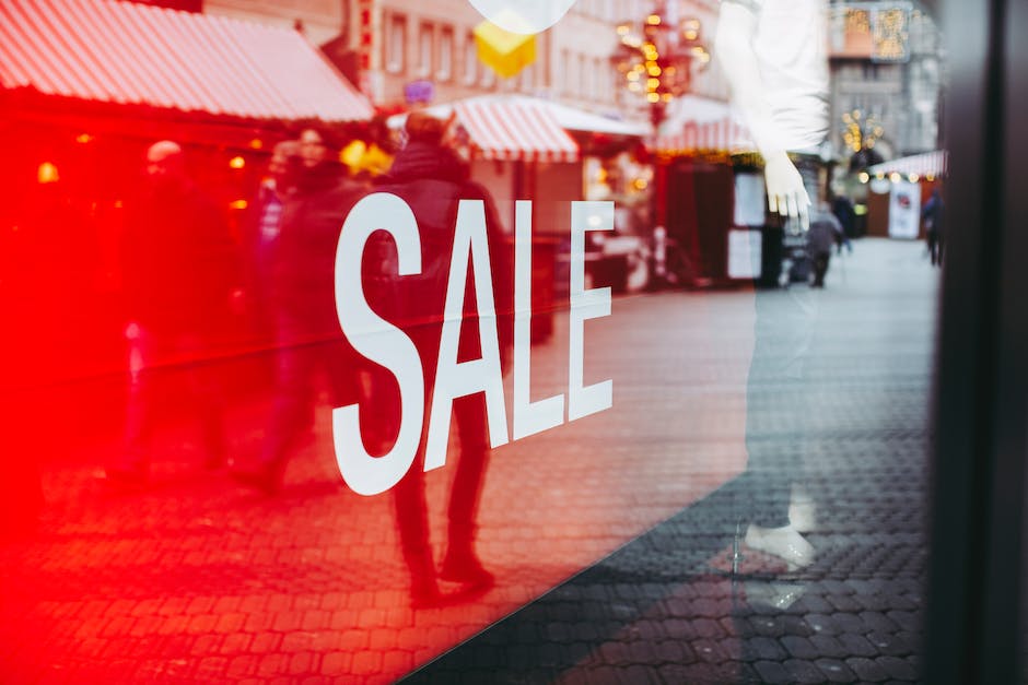 A bright red SALE sign is displayed on the glass window of a storefront, grabbing the attention of passersby. As people walk by outside, the store also shares company news and email marketing tricks to keep customers engaged