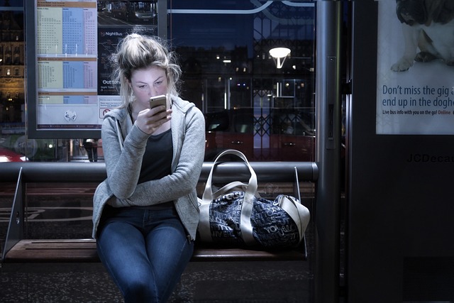 A person sits on a bench at a bus stop, looking down at their smartphone, perhaps reading personalized messaging. A large bag is placed next to them, and the background includes a schedule and a partially visible advertisement.