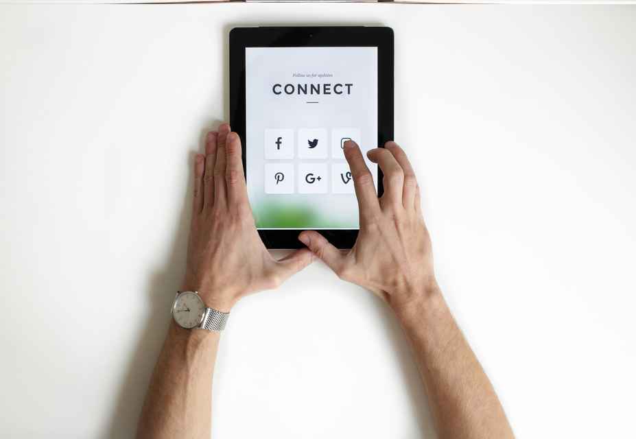 A person uses a tablet displaying a CONNECT screen with various social media icons, including Facebook, Twitter, LinkedIn, Pinterest, Google+, and Vine, effectively managing their social media strategy.