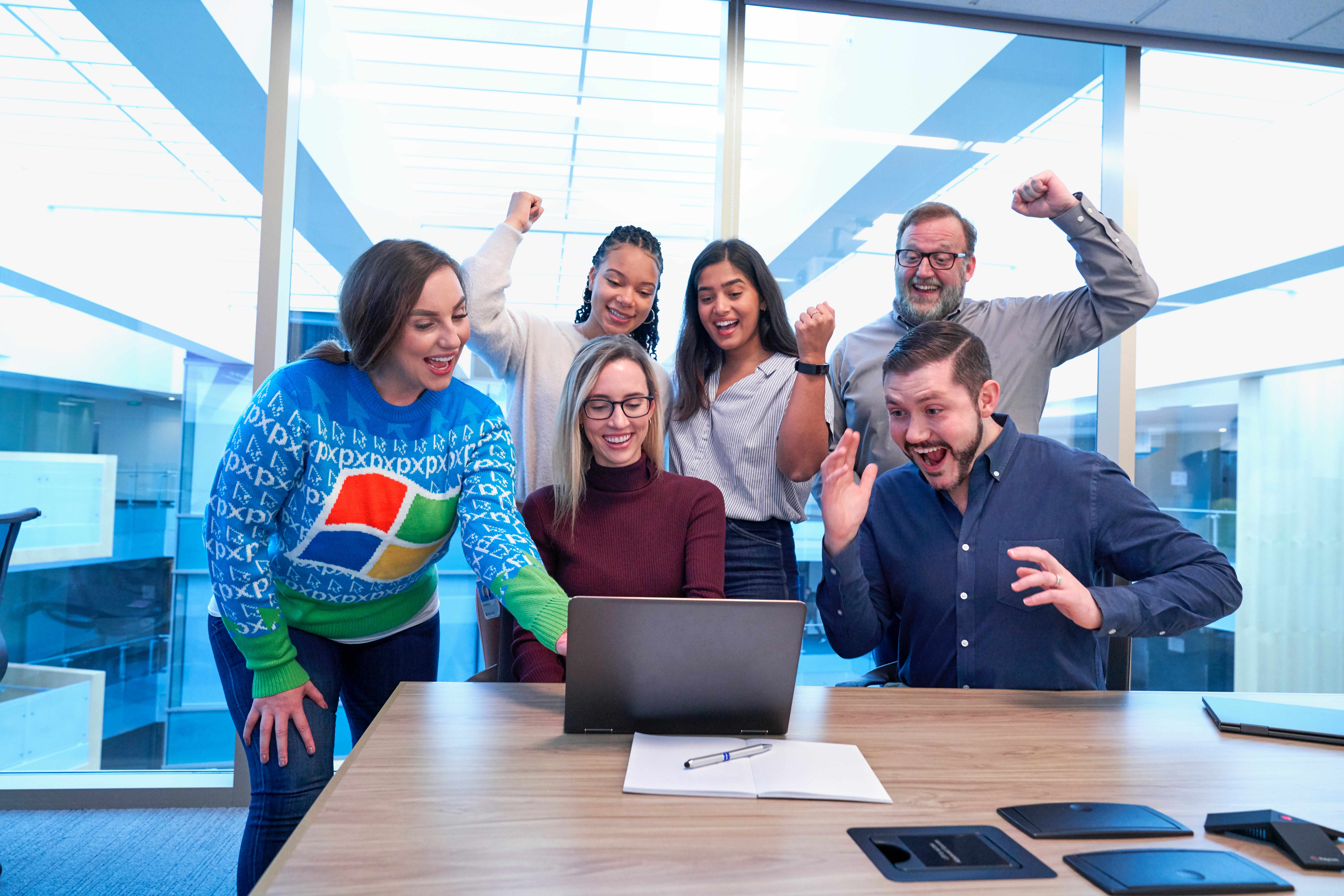 Six people gathered around a laptop in a modern office setting, looking excited and celebrating with raised arms. The group includes three women and three men.