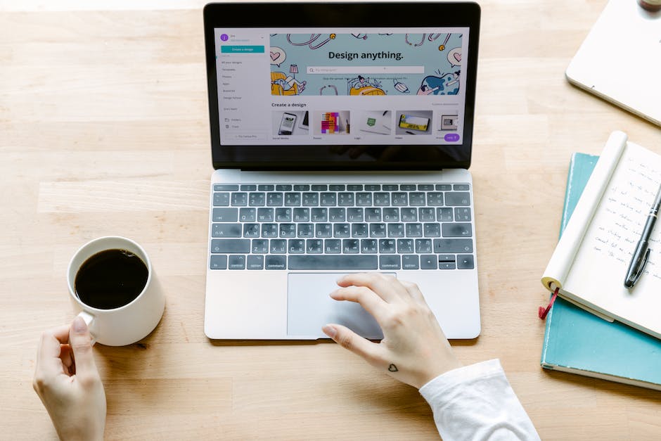 A person with a small heart tattoo on their wrist uses a laptop displaying a design website focused on User Experience (UX), while holding a cup of coffee in their other hand. Notebooks and a pen lie on the wooden table beside them.