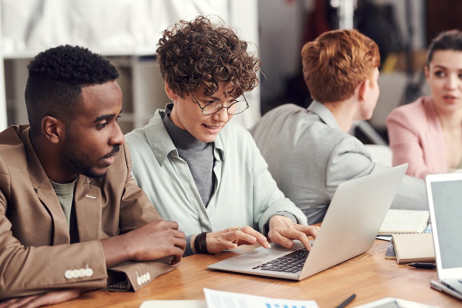 Four people sitting at a table with laptops, engaged in discussion. The focus is on a person with curly hair and glasses looking at a laptop screen discussing affordable web design solutions, while another listens attentively.