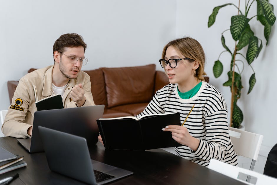 Two people sit at a desk with laptops, discussing something in notebooks. A leafy plant is in the background near a couch. They seem focused on how email marketing can boost your business growth.