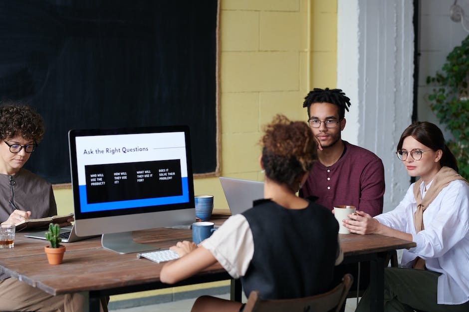 Four people are seated around a table with laptops and notebooks, engaging in a discussion about affordable web design solutions. A monitor displays a slide titled Ask the Right Questions.