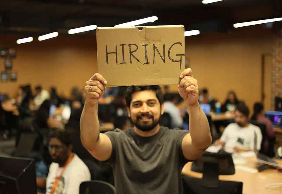 Person holding a cardboard sign that says HIRING in an office with people working at desks in the background, showcasing effective talent attraction strategies.